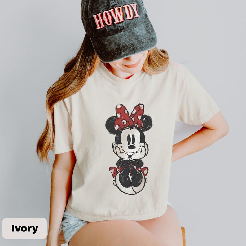 K&ouml;nnte beinhalten: Ein elfenbeinfarbenes Kurzarm-T-Shirt mit einer Grafik einer Zeichentrickmaus mit roter, gepunkteter Schleife, kombiniert mit einer verwaschenen schwarzen Baseballkappe, auf deren Vorderseite das Wort HOWDY in roten Buchstaben gestickt ist.