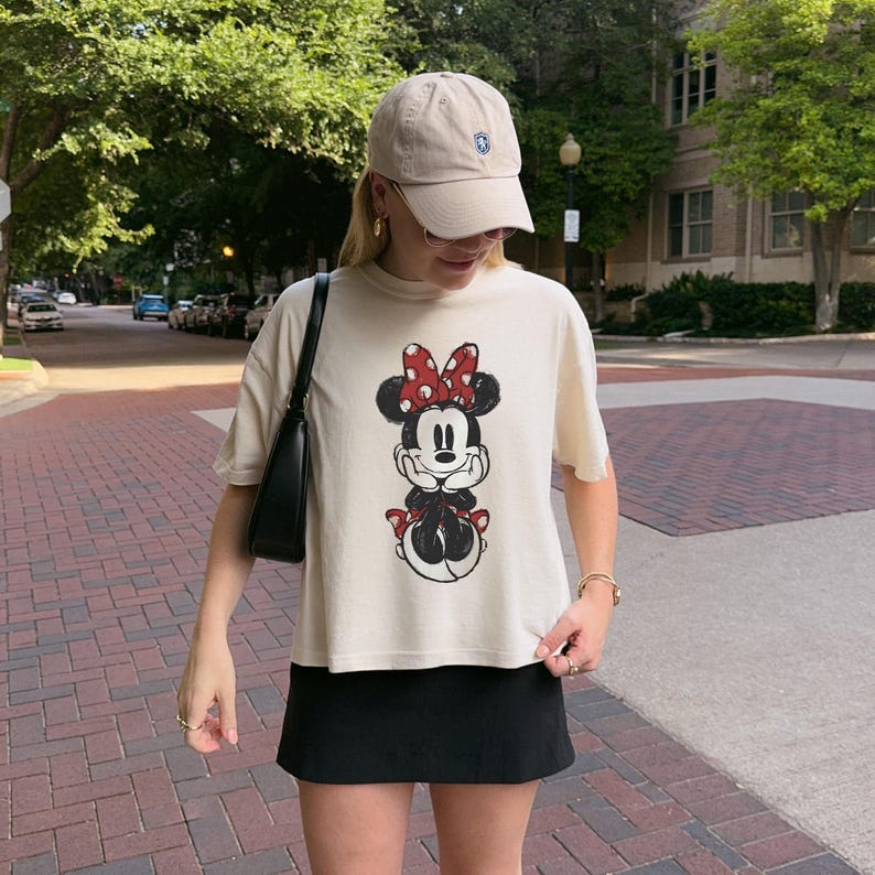 K&ouml;nnte beinhalten: Ein beiges Kurzarm-T-Shirt mit einem Grafikdruck von Minnie Maus mit einer rot-wei&szlig;en gepunkteten Schleife, kombiniert mit einer schwarzen Baseballkappe, einer schwarzen Umh&auml;ngetasche und einem schwarzen Rock. Das T-Shirt hat einen Rundhalsausschnitt.