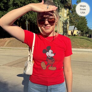 Puede incluir: Una camiseta roja con un dise&ntilde;o de Mickey Mouse. La camiseta tiene mangas cortas y cuello redondo. La persona lleva gafas de sol y un peque&ntilde;o bolso blanco con una cadena dorada.