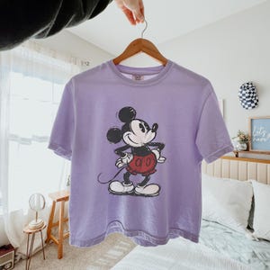 Puede incluir: Una camiseta de manga corta de color morado claro con un gr&aacute;fico de estilo vintage de Mickey Mouse en negro, blanco y rojo. El personaje aparece de pie con las manos en la cintura, en un estampado texturizado y desgastado en el pecho.