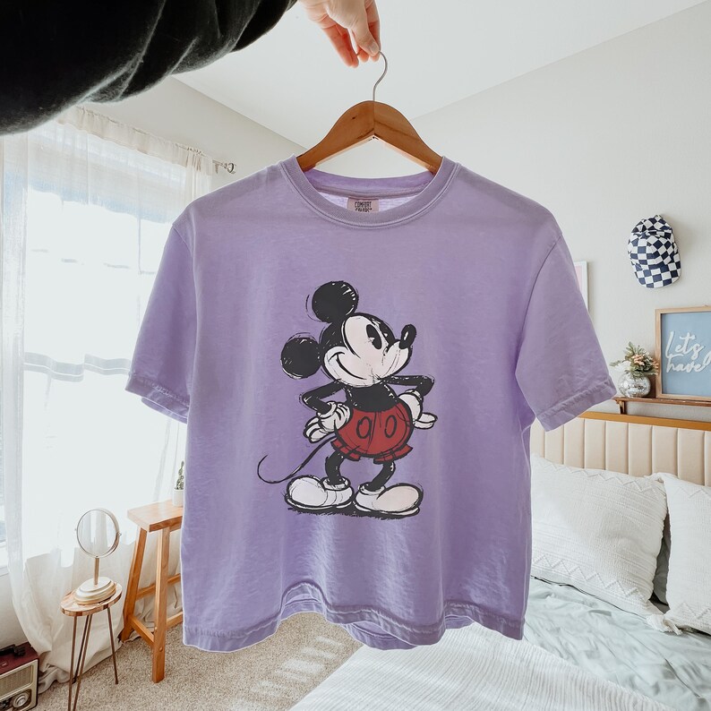 Puede incluir: Una camiseta de manga corta de color morado claro con un gr&aacute;fico de estilo vintage de Mickey Mouse en negro, blanco y rojo. El personaje aparece de pie con las manos en la cintura, en un estampado texturizado y desgastado en el pecho.