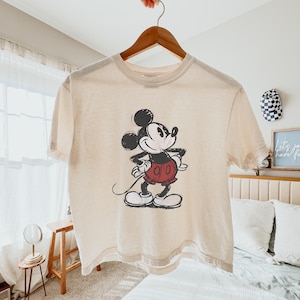 Puede incluir: Una camiseta de manga corta de color crema con un gr&aacute;fico desgastado en blanco y negro de Mickey Mouse con pantalones cortos rojos. El personaje aparece en una pose cl&aacute;sica de pie con las manos en la cintura, centrado en la prenda.