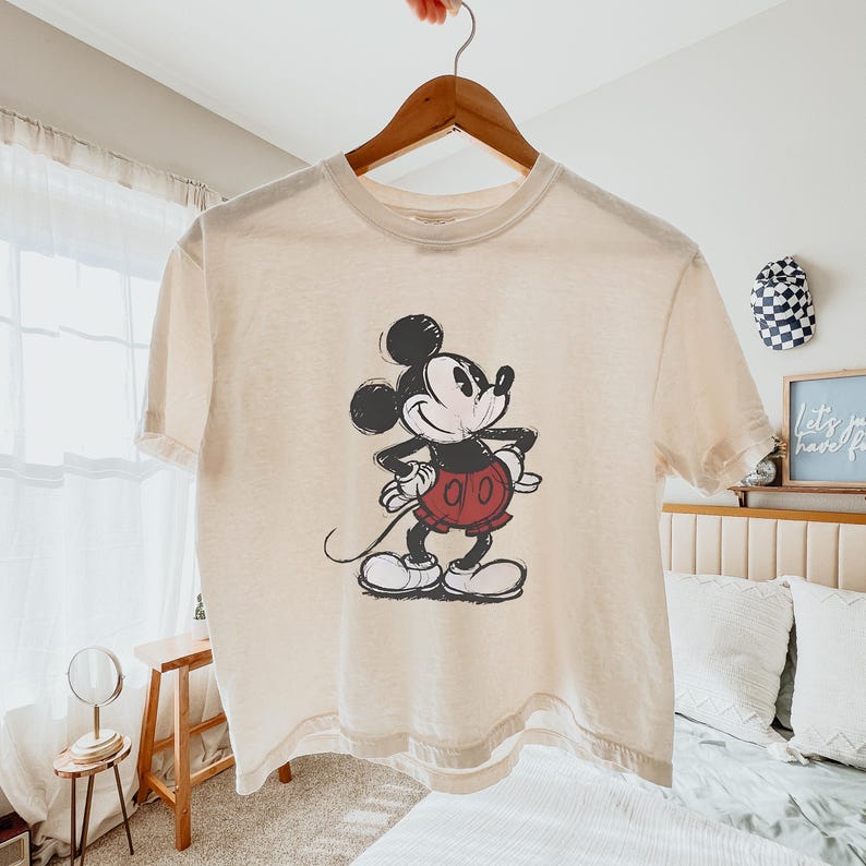 Puede incluir: Una camiseta de manga corta de color crema con un gr&aacute;fico desgastado en blanco y negro de Mickey Mouse con pantalones cortos rojos. El personaje aparece en una pose cl&aacute;sica de pie con las manos en la cintura, centrado en la prenda.