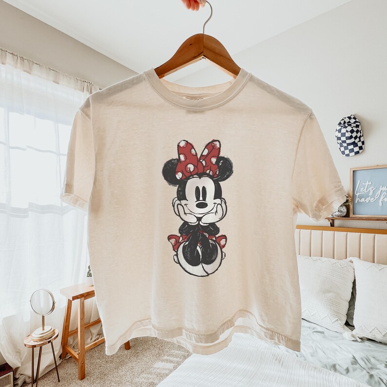K&ouml;nnte beinhalten: Ein cremefarbenes Kurzarm-T-Shirt mit einem verwaschenen Grafikdruck von Minnie Mouse. Die Figur ist mit einer rot-wei&szlig;en gepunkteten Schleife, schwarzen Ohren sowie einem schwarz-wei&szlig;en K&ouml;rper dargestellt und st&uuml;tzt ihr Kinn auf ihre H&auml;nde.