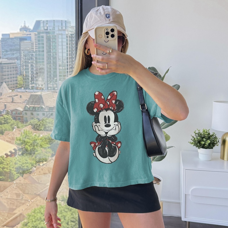 K&ouml;nnte beinhalten: Ein kurz&auml;rmeliges T-Shirt in Blaugr&uuml;n mit einem Grafikdruck von Minnie Maus mit einer rot-wei&szlig; gepunkteten Schleife. Das Shirt wird mit einem schwarzen Rock und einer schwarzen Umh&auml;ngetasche kombiniert. Dazu wird eine beige Baseballkappe getragen.