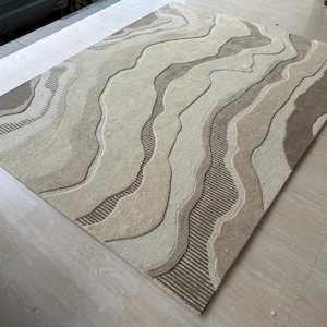 Op de afbeelding: Een rechthoekig vloerkleed met een abstract ontwerp in beige, bruin en bruin. Het vloerkleed heeft een getextureerd, verhoogd patroon dat lijkt op gelaagde, organische vormen. Het ontwerp bevat zowel effen als gestreepte secties.
