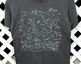 May include: Dark gray t-shirt featuring a detailed constellation map with connecting lines and star names in white and light green. The text "HEAVENLY BODIES" is printed across the bottom of the design. The shirt is hanging on a wooden hanger.