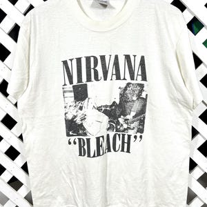 Puede incluir: Camiseta blanca con el nombre de la banda "NIRVANA" en letras mayúsculas negras sobre una imagen en blanco y negro. La palabra "BLEACH" está impresa entre comillas debajo, en negrita.