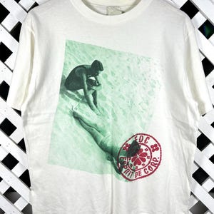 May include: A white short-sleeve t-shirt featuring a green-toned photographic print of two figures on a sandy beach. A circular red stamp graphic is overlaid on the print, containing the text EDC ESPRIT DE CORP. and a floral icon.