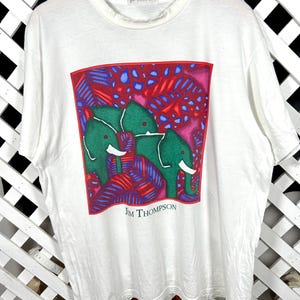 May include: A white short-sleeved t-shirt featuring a square graphic print of two green elephants surrounded by red and blue foliage. The text "JIM THOMPSON" is printed in black below the graphic and on the inner neck label of the shirt.
