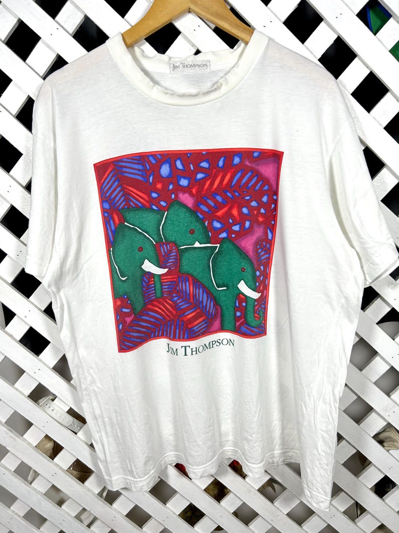 May include: A white short-sleeved t-shirt featuring a square graphic print of two green elephants surrounded by red and blue foliage. The text "JIM THOMPSON" is printed in black below the graphic and on the inner neck label of the shirt.