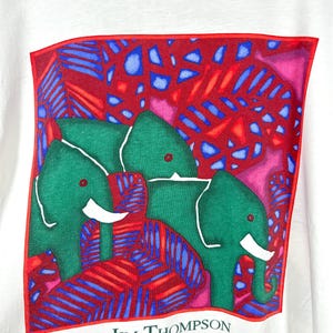 May include: A white t-shirt featuring a square graphic print of two green elephants with white tusks set against a vibrant background of red and blue tropical leaves. The name JIM THOMPSON is printed in black text below the square graphic.