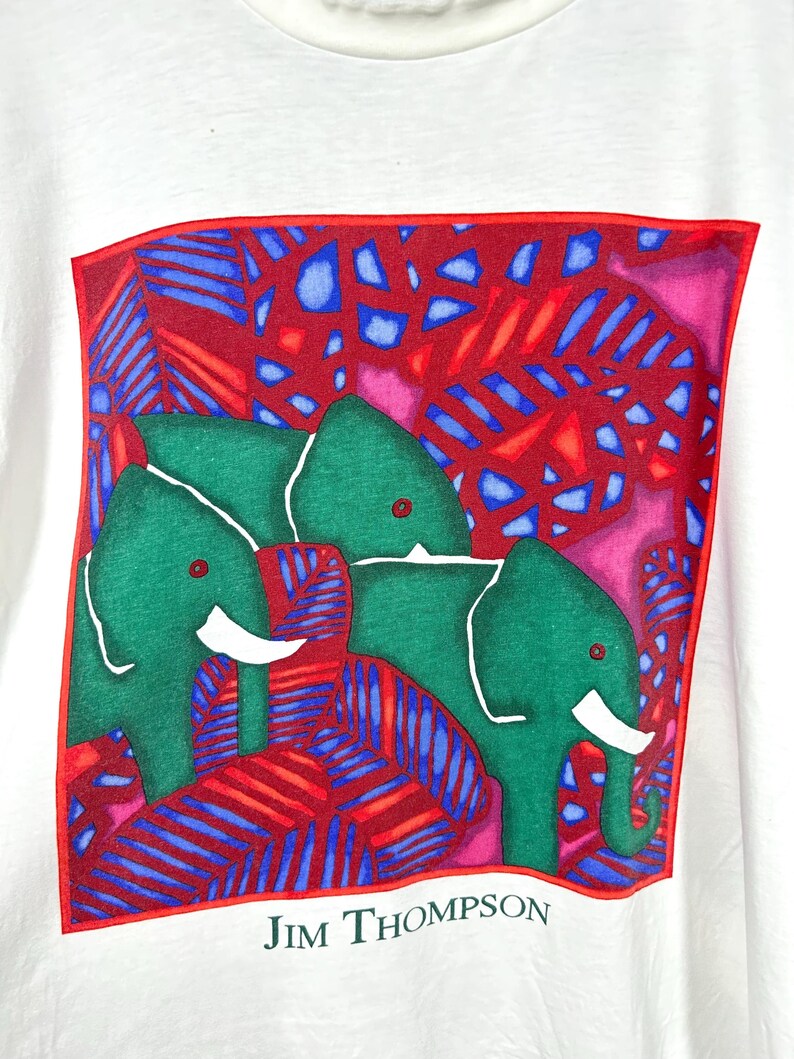May include: A white t-shirt featuring a square graphic print of two green elephants with white tusks set against a vibrant background of red and blue tropical leaves. The name JIM THOMPSON is printed in black text below the square graphic.