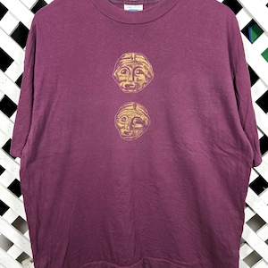 Peut inclure: Un t-shirt de couleur bordeaux avec deux graphiques de visage stylisés de couleur or. Le t-shirt a un col rond et des manches courtes. Le t-shirt est suspendu à un cintre en bois sur un fond de treillis blanc.