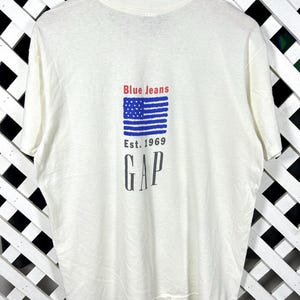 Peut inclure: Un t-shirt blanc à manches courtes avec un motif au dos. Le motif comprend le texte rouge Blue Jeans, un drapeau américain stylisé bleu, le texte Est. 1969 et le mot GAP en grandes lettres capitales noires centré sur le tissu.