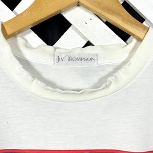 May include: A white t-shirt with a ribbed crew neckline and a label reading Jim Thompson. The shirt features a graphic print with a red border, containing abstract shapes in shades of blue, purple, and red. The garment hangs on a wooden hanger.