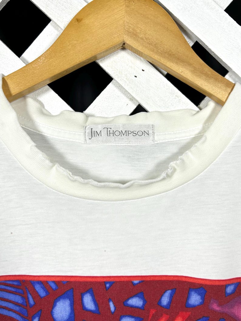 May include: A white t-shirt with a ribbed crew neckline and a label reading Jim Thompson. The shirt features a graphic print with a red border, containing abstract shapes in shades of blue, purple, and red. The garment hangs on a wooden hanger.