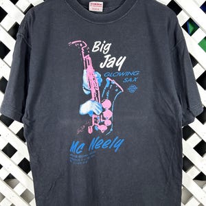 May include: A black short-sleeve t-shirt featuring a graphic of a pink saxophone being played by white-gloved hands. The text reads Big Jay GLOWING SAX Mc Neely, with additional small text below. The shirt has a crew neckline and a standard fit.