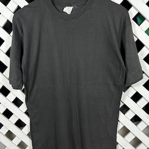 Könnte beinhalten: Ein anthrazitfarbenes Kurzarm-T-Shirt hängt an einem Holzbügel. Das Shirt hat einen Rundhalsausschnitt und ein Etikett ist in der Nähe des Kragens sichtbar. Der Hintergrund ist ein weißes Gitter.