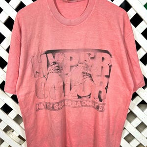 May include: A faded pink t-shirt with a crew neck and short sleeves. The front features a black graphic with the word "HYPERCOLOR" and the text "NYE GENERATION" in a film strip design. The shirt is hanging on a wooden hanger.