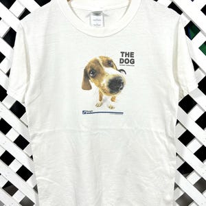 May include: White t-shirt featuring a close-up image of a beagle with the text "THE DOG" above the image and "Artist Collection" below. The t-shirt is hanging on a wooden hanger.