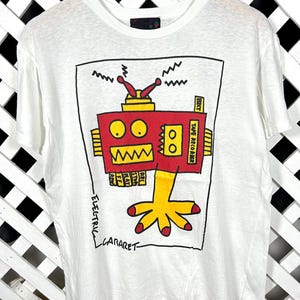May include: A white short-sleeve t-shirt featuring a graphic print of a red robot with yellow accents. The robot has a yellow hand, antennas, and text labels including "TAPE RECORDER" and "ELECTRIC CABARET" in black ink on the front.