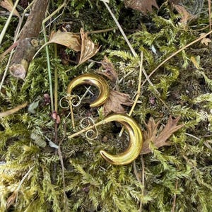 Könnte beinhalten: Ein Paar goldfarbene Creolen mit einem dicken, halbmondförmigen Design. Jeder Ohrring verfügt über eine polierte, reflektierende Oberfläche und einen Standard-Steckerverschluss mit Schmetterlingsmutter, die eine glatte, geschwungene Metallstruktur aufweisen.