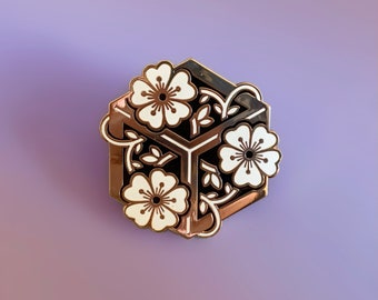May include: A gold-toned enamel pin featuring a geometric design with three white flowers and black leaves. The flowers are stylized and have a simple design.