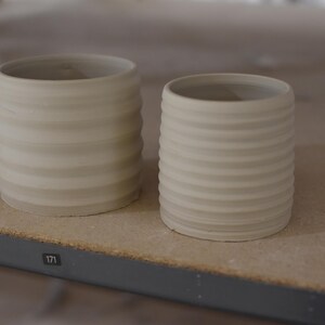 May include: Two white ceramic cups with a ribbed texture. The cups are sitting on a wooden surface.