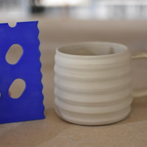 May include: A blue plastic tool with two oval-shaped holes, used for smoothing and shaping clay, next to a white ceramic mug with a wavy design.