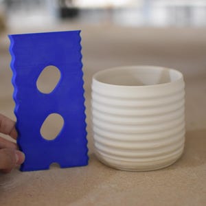 May include: A white ceramic mug with a ribbed design, next to a blue plastic scraper with two oval-shaped holes.