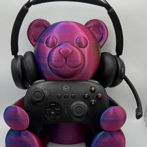May include: A 3D-printed bear-shaped stand in a metallic purple and blue gradient finish, holding a black video game controller. A pair of black over-ear headphones with a microphone boom is resting on the head of the bear figure.