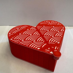May include: A red heart-shaped box with a white wave pattern on the lid. The box has a small, round knob on the side. The box is made of a textured material.