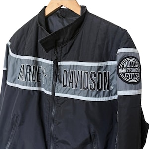 May include: Black Harley-Davidson motorcycle jacket with a gray stripe across the chest and the brand name in large white letters. The jacket has a zip-up front, a stand-up collar, and a Harley-Davidson patch on the sleeve.