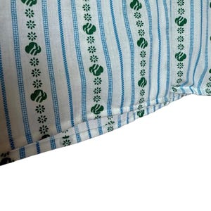 May include: Close-up of white fabric featuring a repeating pattern of green floral and cloud-like shapes, and blue vertical stripes. The fabric is folded at the bottom, showing the edge and stitching.