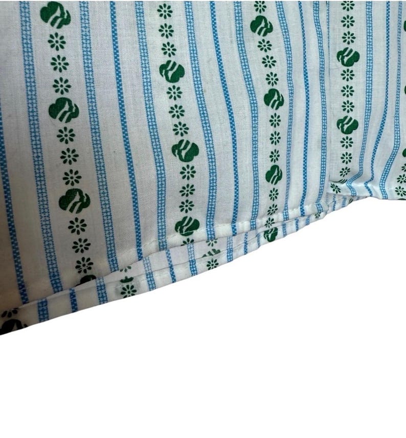 May include: Close-up of white fabric featuring a repeating pattern of green floral and cloud-like shapes, and blue vertical stripes. The fabric is folded at the bottom, showing the edge and stitching.