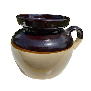 Puede incluir: Un tarro de gres vintage con una parte superior vidriada de color marrón oscuro y una base de color crema. El tarro presenta un cuerpo redondeado, un asa y una banda marrón. Esta pieza decorativa es ideal para la cocina o la decoración del hogar.