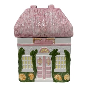 Peut inclure: Un pot à biscuits en céramique en forme de cottage avec un toit de chaume rose texturé. La maison blanche présente une porte rose centrale, des fenêtres à croisillons jaunes, des vignes vertes et des fleurs orange à la base.
