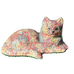 May include: A ceramic cat ornament in a reclining position, featuring a vibrant paisley design in red, yellow, and blue. The cat has black eyes, and the base is trimmed with green. A decorative item, suitable for display.