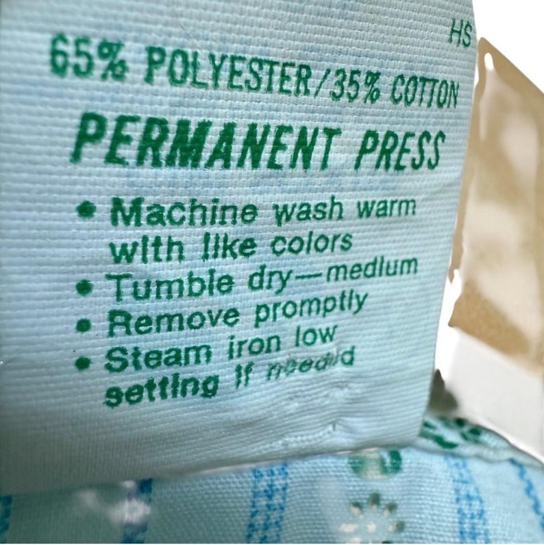 May include: A close-up of a light blue fabric label with green text. The label states "65% POLYESTER/35% COTTON" and "PERMANENT PRESS." Washing instructions and care details are also printed on the label.