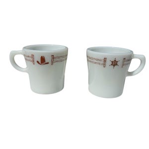 May include: Two white ceramic mugs with brown cowboy-themed designs. One mug features a cowboy hat, and the other has a sheriff's star. Both mugs have a brown fence design around the top.