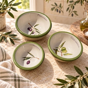 May include: Three round ceramic bowls with a cream-colored interior and a green rim. Each bowl features a painted olive branch illustration and the text "Provençal," "Niçoise," or "Cerignola" printed along the inner edge of the bowl rim.