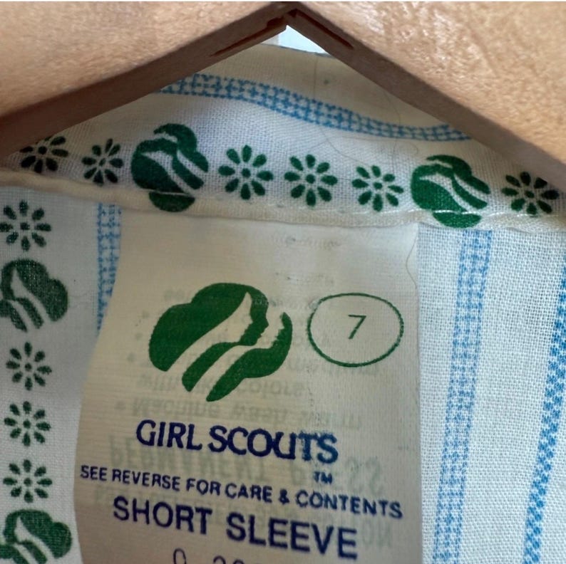 May include: Close-up of a Girl Scouts short sleeve shirt label. The label is white with green and blue details. The Girl Scouts logo is visible, along with the number 7. The shirt has a blue and white striped pattern.