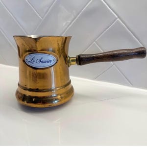 May include: A vintage copper coffee pot with a wooden handle. The pot has a rounded base and a flared top. A small, oval-shaped label on the side reads "Le Saucier". The handle is dark brown, and the pot has a warm, aged patina.