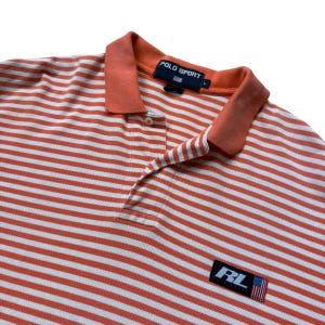 May include: Orange and white striped Polo Sport shirt with a collared neckline. The shirt features a small patch with the letters "RL" and an American flag. The shirt has a buttoned placket and a "POLO SPORT" label.