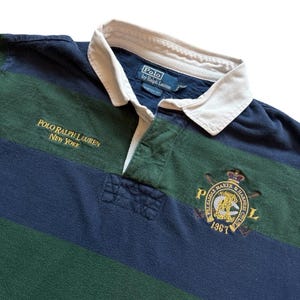 May include: A navy blue, green, and white striped rugby shirt. The shirt features the text "POLO RALPH LAUREN NEW YORK" in gold lettering. A crest with the text "PRL SADDLE MAKER & HARNESS 1967" is embroidered on the chest.