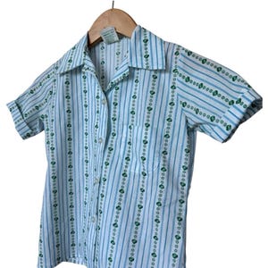May include: A short-sleeved shirt with a light blue and white striped design. The shirt has vertical rows of green floral patterns and a small chest pocket. The collar and cuffs are folded over.