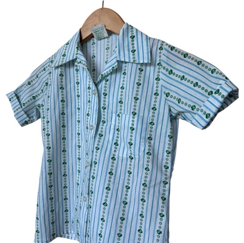 May include: A short-sleeved shirt with a light blue and white striped design. The shirt has vertical rows of green floral patterns and a small chest pocket. The collar and cuffs are folded over.