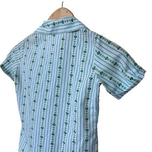 May include: A light blue and white short-sleeved shirt with vertical stripes and green heart and flower patterns. The shirt is hanging on a wooden hanger.