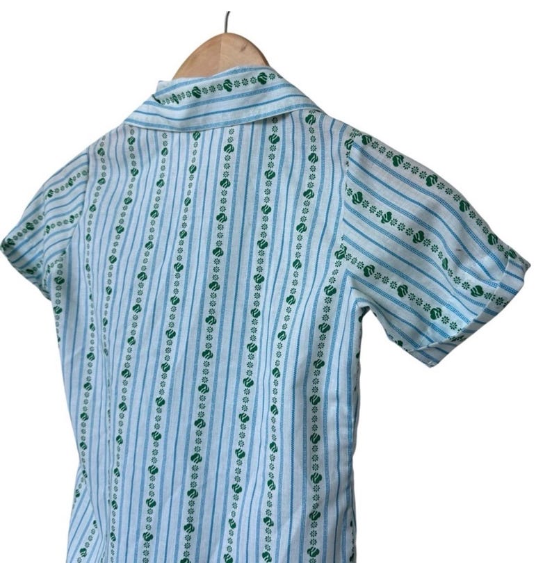 May include: A light blue and white short-sleeved shirt with vertical stripes and green heart and flower patterns. The shirt is hanging on a wooden hanger.